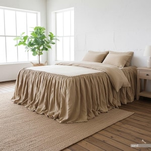 May include: A bed with a beige linen bedspread and matching pillows. The bed has a ruffled bed skirt and is set on a woven jute rug. A potted plant sits near a window, and a nightstand with a lamp is visible.