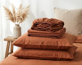 Può includere: Un set di biancheria da letto color ruggine che include due federe rettangolari, un lenzuolo piano piegato e un lenzuolo con angoli impilati sopra un copriletto abbinato. Il tessuto ha una trama visibile in una tonalità terracotta.