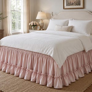 May include: A bed featuring a light pink bed skirt with a gathered ruffle design along the bottom edge. The bed is dressed in a white textured quilt, two white pillows, and a small rectangular accent pillow placed in the center of the bed.