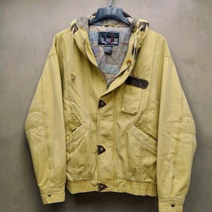 Pode incluir: Um casaco com capuz amarelo-mostarda com fecho de correr frontal, três detalhes triangulares em imitação de pele castanha, um bolso no peito com aba castanha e um forro interior estampado com o texto Performance na etiqueta interna.
