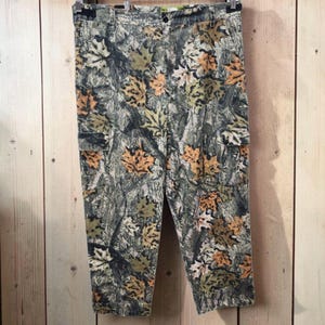 Pode incluir: Calças cargo com um padrão de camuflagem de folhas de ácer de outono em tons de castanho, verde e bege sobre um fundo de casca de árvore texturizada. As calças incluem fecho de botão, presilhas para cinto e dois bolsos cargo laterais.