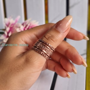 May include: A copper-toned metal ring featuring a stacked design with three distinct bands. The central band displays an embossed clover-like pattern, while the outer bands have a textured finish. The text Elarvienne Jewels is visible.