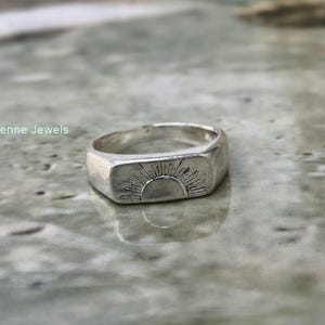 May include: A silver signet ring featuring an engraved sunburst design on the flat rectangular face. The band is smooth and polished, with the text Elarvienne Jewels visible in the upper left corner of the frame.