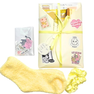 May include: A gift set with a yellow theme, including a wrapped package tied with a ribbon, stickers, a pair of fuzzy socks, and a scrunchie. The package has a tag that says "Comic Issue 1-5". Stickers say "Rather be reading" and "Let me read in peace".