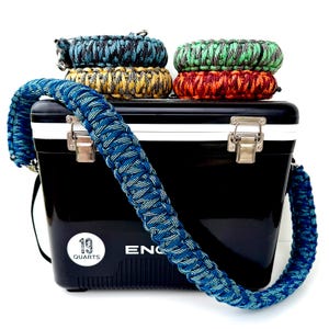 May include: A black cooler with a blue and white braided strap and handle. The cooler has a white trim and silver latches. Several braided bracelets in various colors, including blue, green, yellow, and red, are stacked on top. The cooler is labeled "19 Quarts".