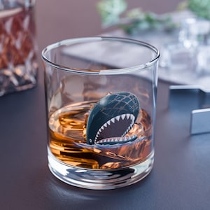 May include: A clear, round whiskey glass filled with amber liquid and ice cubes. A shark-shaped ice cube with a dark blue body and white teeth floats in the drink. The glass sits on a dark surface.
