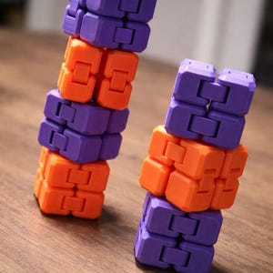 May include: Two fidget toys, each made of interlocking blocks in purple and orange. The blocks are stacked to form a tower-like structure. The toys are on a wooden surface.