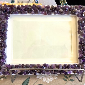 May include: A rectangular white picture frame featuring a border densely covered in small, polished purple amethyst stones. The frame is supported by a silver-toned metal stand and contains a plain, light-colored insert in the center.