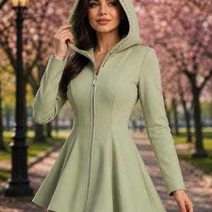 May include: A pale green hooded coat with a fitted bodice and flared skirt. The coat has a full-length zip and decorative stitching on the hood. The coat is worn outdoors in a park setting, with a backdrop of trees.