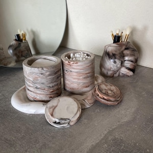 May include: A marble-effect bathroom set comprising a tray, two cylindrical containers, a unique vase, and a small round container. The set has a swirling pattern of white, brown, and pink tones. The containers hold cotton pads, cotton buds, and makeup brushes.