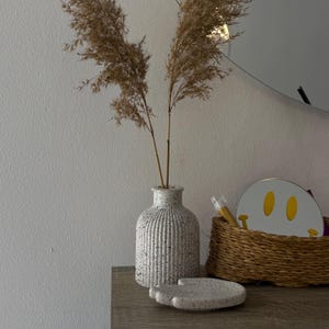 May include: A decorative arrangement featuring a speckled white vase with vertical ridges, holding dried brown reeds. A small, speckled tray and a woven basket with a smiley face mirror are also present, creating a minimalist aesthetic.