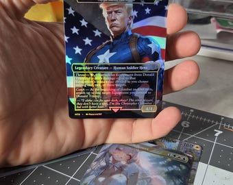 以下が含まれることがあります： アメリカ国旗を背景にキャプテン・アメリカ風の兵士として描かれたドナルド・トランプのホログラフィック・トレーディングカード。カードには「Donald Trump, US. President」、「Legendary Creature - Human Soldier Hero」、パワーとタフネス「4/4」のテキストが記載されています。