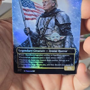 May include: A collectible trading card featuring a fantasy illustration of a figure holding an American flag. The card is titled "Trump, the Dark Lord" and includes the text "Legendary Creature - Avatar Horror". The card has a holographic finish.