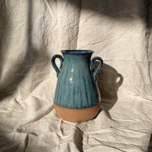 Könnte beinhalten: Eine Keramikvase mit zwei Henkeln, einer glänzend blau glasierten oberen Hälfte und einem naturbelassenen, unglasierten Terrakotta-Fuß. Das Gefäß besitzt eine weite Öffnung und einen bauchigen Körper, der einen deutlichen Kontrast aufweist.