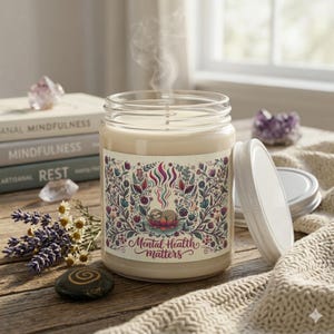 May include: A lit candle in a clear glass jar with a white lid. The candle has a label with a floral design and the text "Mental Health Matters". The candle is on a wooden surface with books and crystals.