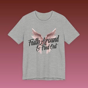 May include: A heather gray t-shirt featuring a design of angel wings in shades of pink and white. The text "Faith Around & Find Out" is printed in black script below the wings. The shirt has a crew neck and short sleeves.