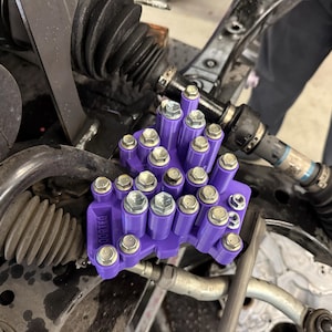 May include: A purple plastic bolt organizer tray featuring multiple cylindrical slots holding various silver-colored hexagonal bolts, with the word "DORTEO" embossed on the side, positioned within a vehicle engine bay.