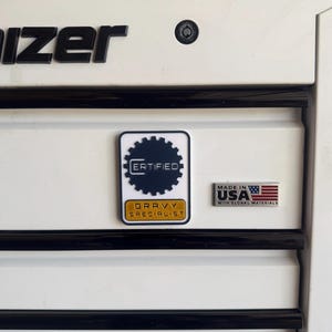 May include: A white metal tool chest drawer featuring a rectangular sticker with a dark blue gear icon, the text CERTIFIED, and a gold bar with the text GRAVY SPECIALIST. Next to it is a small rectangular metal badge reading MADE IN USA WITH GLOBAL MATERIALS.