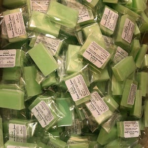 May include: A pile of light green soap bars, each individually wrapped in clear plastic. The rectangular bars have small white labels with black text. The soaps are stacked together, creating a full frame image.