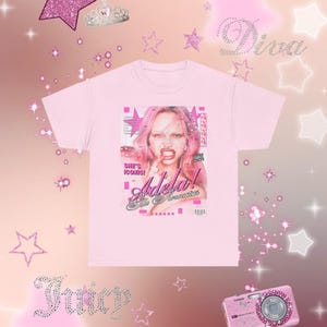Puede incluir: Una camiseta rosa claro con un gráfico estilo revista de una mujer con cabello rosado. El gráfico incluye el texto "She's Iconic! Adela! The Reincarnation". El fondo está decorado con estrellas rosas y las palabras "Diva" y "Juicy".