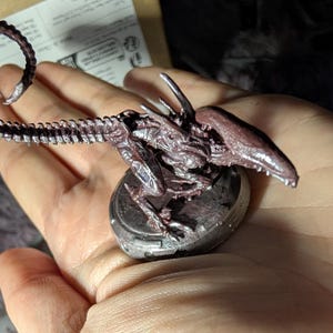 May include: A detailed miniature figurine of a Xenomorph, featuring a dark purple and silver color scheme. The creature has a long, segmented tail and is mounted on a circular base. The figurine's design includes intricate details.