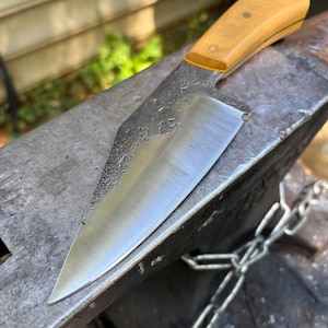 May include: A fixed-blade knife with a polished, sharp steel edge and a textured, dark forged finish on the spine. The handle is made of light-colored wood, secured with three metal pins, and features a thin yellow accent layer near the blade.