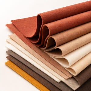 May include: A stack of rectangular felt sheets in a gradient of warm earth tones, including shades of deep rust, terracotta, tan, cream, beige, dark brown, and mustard yellow. The sheets are layered and folded to show the soft, matte texture.