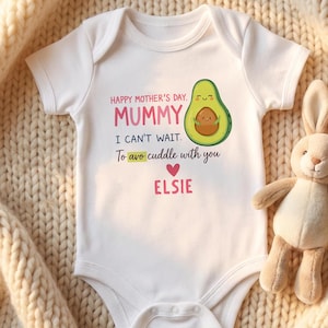 May include: White baby onesie with the text "HAPPY MOTHER'S DAY, MUMMY, I CAN'T WAIT. To avo cuddle with you, ELSIE" and a cartoon avocado illustration. A stuffed bunny is next to the onesie on a cream-colored knitted blanket.