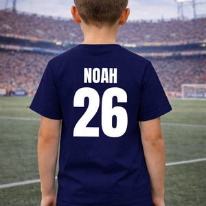 Puede incluir: Una camiseta azul marino con el nombre "NOAH" y el n&uacute;mero "26" impresos en blanco en la espalda. La camiseta la lleva una persona de pie en un campo de f&uacute;tbol.