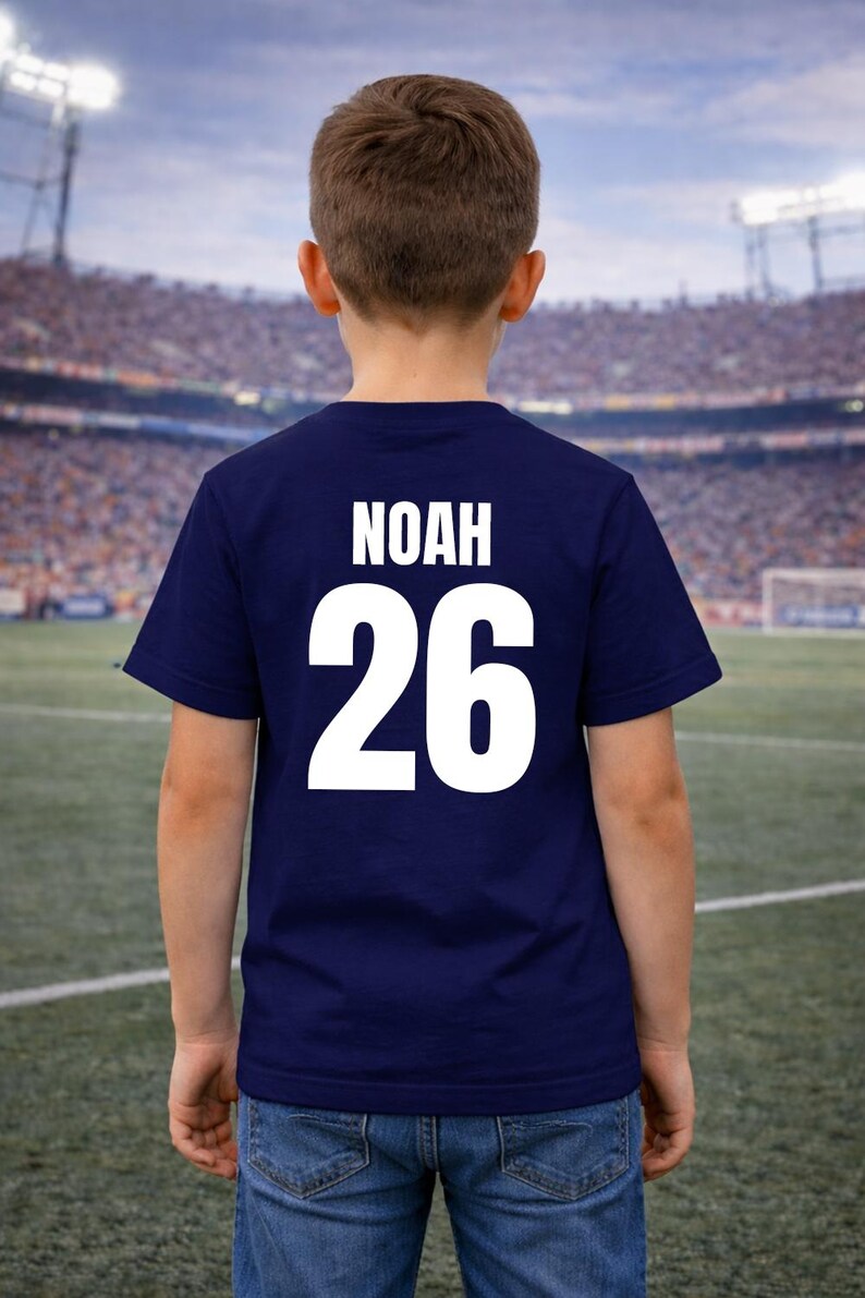 Puede incluir: Una camiseta azul marino con el nombre "NOAH" y el n&uacute;mero "26" impresos en blanco en la espalda. La camiseta la lleva una persona de pie en un campo de f&uacute;tbol.