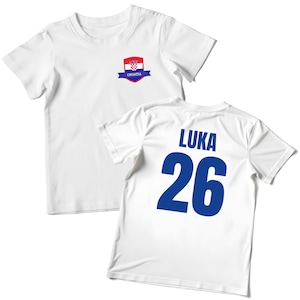 K&ouml;nnte beinhalten: Wei&szlig;es T-Shirt mit einem kroatischen Flaggenemblem auf der Vorderseite. Auf der R&uuml;ckseite des T-Shirts steht der Name "LUKA" &uuml;ber der Nummer "26" in Blau. Das T-Shirt besteht aus leichtem Material.
