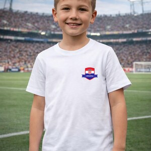 K&ouml;nnte beinhalten: Ein wei&szlig;es T-Shirt mit einem kleinen kroatischen Flaggenemblem auf der Brust. Das Emblem zeigt das kroatische Wappen und das Wort "CROATIA" auf einem blauen Band. Das T-Shirt wird von einer jungen Person vor einem Stadion getragen.