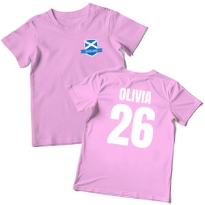 Puede incluir: Dos camisetas de color rosa claro. Una camiseta presenta un peque&ntilde;o emblema con la bandera escocesa y la palabra "Scotland". La otra camiseta muestra el nombre "Olivia" y el n&uacute;mero "26" en blanco.