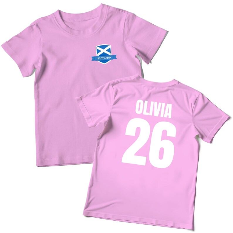 Puede incluir: Dos camisetas de color rosa claro. Una camiseta presenta un peque&ntilde;o emblema con la bandera escocesa y la palabra "Scotland". La otra camiseta muestra el nombre "Olivia" y el n&uacute;mero "26" en blanco.