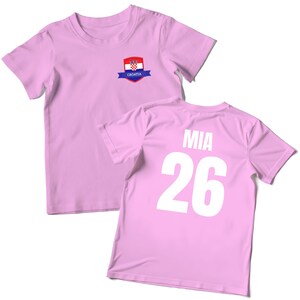 K&ouml;nnte beinhalten: Zwei hellrosa T-Shirts. Eines hat ein kroatisches Flaggenemblem auf der Vorderseite. Das andere hat den Namen "MIA" und die Nummer "26" in Wei&szlig; auf der R&uuml;ckseite aufgedruckt. Die Shirts sind aus weichem Material.