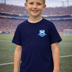 Puede incluir: Camiseta azul marino con un escudo de Escocia en el pecho. El escudo incluye una X blanca sobre fondo azul con una pancarta que dice "Scotland". La camiseta la lleva un joven frente a un estadio.