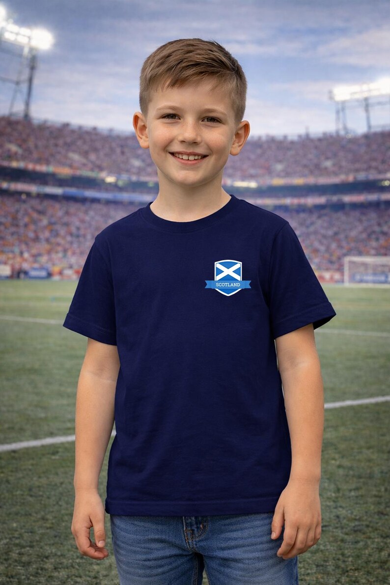 Puede incluir: Camiseta azul marino con un escudo de Escocia en el pecho. El escudo incluye una X blanca sobre fondo azul con una pancarta que dice "Scotland". La camiseta la lleva un joven frente a un estadio.