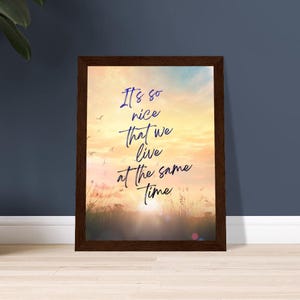 Könnte beinhalten: Gerahmter Druck mit einem Sonnenuntergangs-Landschaftshintergrund. Das Kunstwerk zeigt den Text "It's so nice that we live at the same time" in handschriftlichem Stil. Der Rahmen ist dunkelbraun, und der Druck befindet sich auf einem hellen Holzboden.