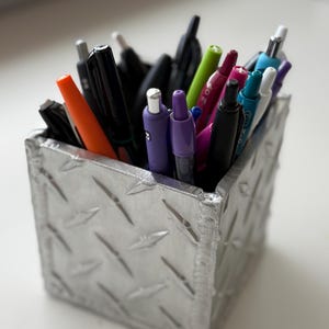 Op de afbeelding: Een zilveren metalen pennenhouder met een ruitpatroon, gevuld met pennen en stiften. De pennen zijn in verschillende kleuren, waaronder zwart, oranje, groen, paars, roze en blauw.