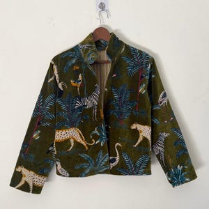 Puede incluir: Chaqueta verde oliva con estampado de jungla. El diseño presenta leopardos, cebras, aves y plantas tropicales. La chaqueta tiene cuello mandarín y mangas largas. Cuelga de una percha de madera.