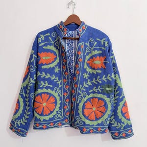 Könnte beinhalten: Eine blaue Jacke mit leuchtend orangefarbenen und hellgrünen Blumenstickereien auf der Vorderseite und den Ärmeln. Das Kleidungsstück hat einen Stehkragen, lange Ärmel und eine offene Vorderseite, die ein weißes Innenfutter mit blauem Blumenmuster zeigt.