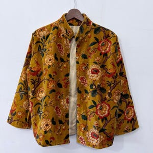 Puede incluir: Una chaqueta de color amarillo mostaza con un estampado de flores y pájaros en tonos rojos, naranjas y verdes. La chaqueta tiene cuello mao, mangas largas y un diseño frontal abierto que revela un forro de rayas verticales blancas y beige.