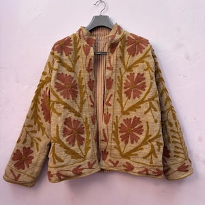 Puede incluir: Una chaqueta beige abierta por delante con bordados texturizados en tonos rosa empolvado y amarillo mostaza. La prenda presenta un patrón floral repetitivo en los paneles frontales y las mangas, con cuello alto y forro interior a rayas.