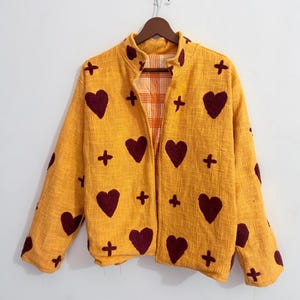 Könnte beinhalten: Eine senfgelbe Jacke mit strukturierter Stoffoberfläche, verziert mit einem wiederkehrenden Muster aus dunkelroten Herzen und kleinen Pluszeichen. Die Jacke hat einen Stehkragen, lange Ärmel und eine offene Vorderseite mit orange-weißem Karofutter.