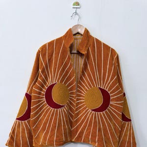Könnte beinhalten: Eine orangefarbene Jacke mit gestickten Sonnen- und Mondsichelmotiven in Gelb und Dunkelrot. Das Design umfasst strahlenförmige, weiße Sticklinien, die von den Himmelskörpern über die Vorderseite und die Ärmel verlaufen, mit einem Stehkragen.