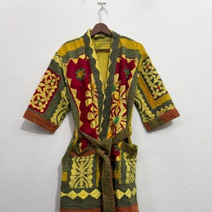 Könnte beinhalten: Ein langärmeliger Morgenmantel mit Patchwork-Design in Gelb, Olivgrün und Dunkelrot. Das Kleidungsstück verfügt über einen Bindegürtel, zwei Vordertaschen und aufwendige Steppmuster auf dem gesamten Stoff und hängt an einem Holzkleiderbügel.