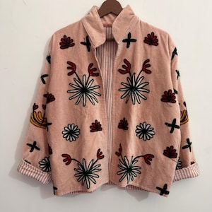 Könnte beinhalten: Eine hellrosa Cordjacke mit offener Vorderseite, Stehkragen und langen Ärmeln mit umgeschlagenen Manschetten. Die Jacke ist mit gestickten Blumenmotiven in Schwarz, Weiß und Rot verziert und hat ein gestreiftes Innenfutter an den Ärmelbündchen.