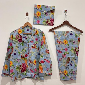 Puede incluir: Un conjunto de pijama azul claro con estampado floral y de pájaros. El conjunto incluye una camisa con botones, pantalones y una bolsa a juego. El ribete es rojo y el diseño presenta flores y pájaros en tonos rojos, amarillos y verdes.