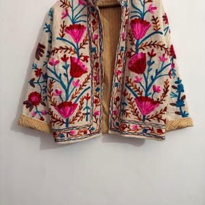 May include: A cream-colored jacket featuring vibrant floral embroidery in shades of hot pink, red, and bright blue. The garment has long sleeves with yellow and white striped cuffs, a high collar, and an interior lining with yellow and white stripes.