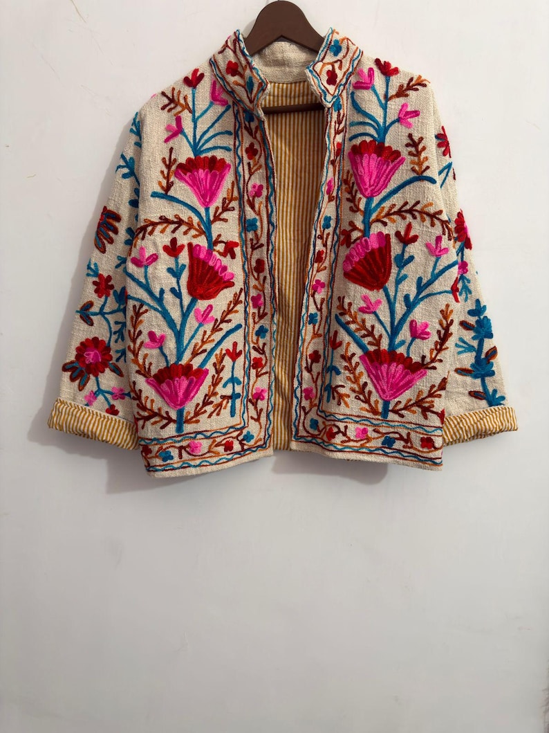 May include: A cream-colored jacket featuring vibrant floral embroidery in shades of hot pink, red, and bright blue. The garment has long sleeves with yellow and white striped cuffs, a high collar, and an interior lining with yellow and white stripes.